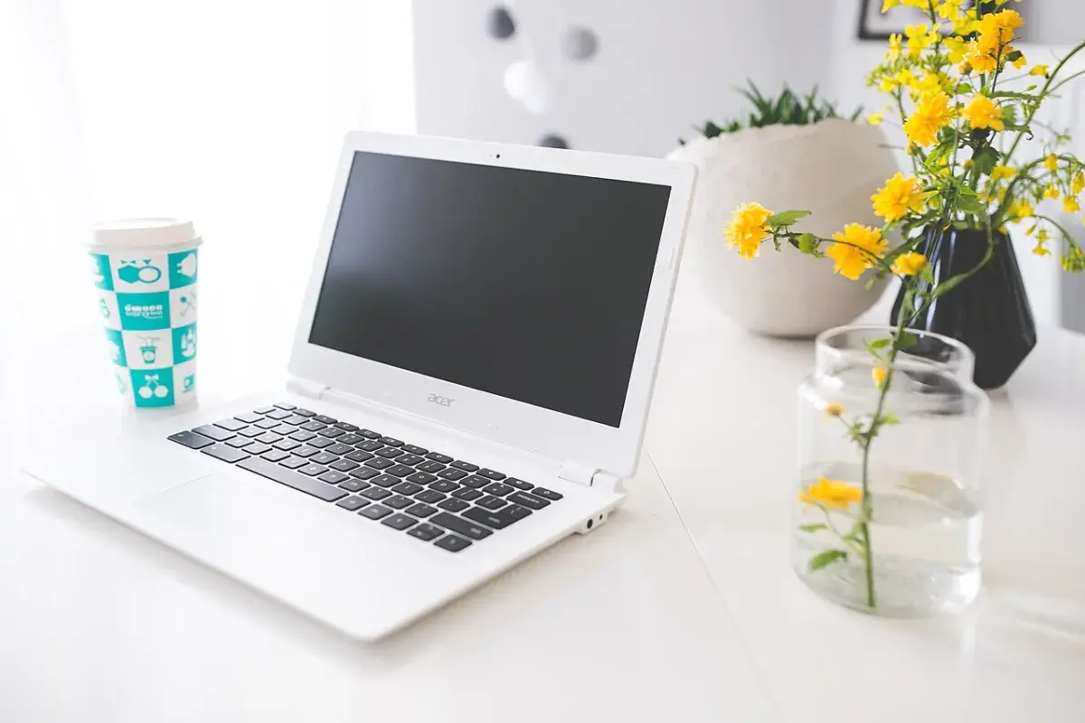 Portátil blanco junto a una bebida y flores amarillas sobre una mesa